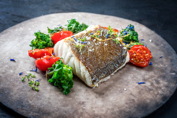Gourmet fried European skrei cod fish filet with kalette and tomatoes as closeup on a modern design plate