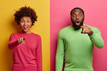 Excited young Afro American woman and man indicate in front, point at camera with surprised...