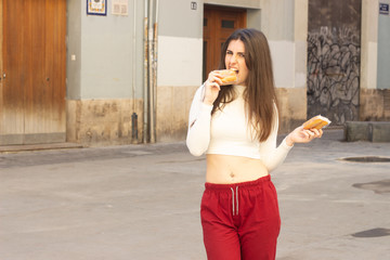Portrait of attractive young woman biting cupcake, lovely smiling, in the city, white top and red pants, dark air. Place for your text in copy space.