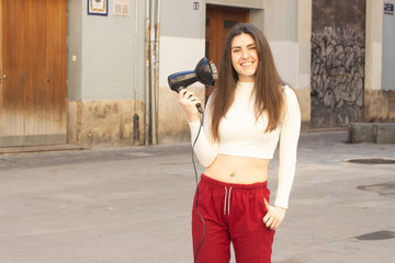 Portrait of attractive caucasian young woman model holding hair dryer, in the city, white top and red pants. Place for your text in copy space.