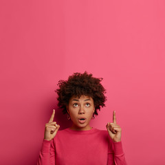 Vertical shot of embarrassed shocked Afro American woman indicates index fingers upwards, impressed by something stunning, shows blank space on pink studio background, stands speechless indoor