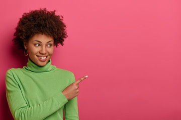 Horizontal shot of smiling curly haired woman indicates at free space, demonstrates place for your advertisement, attracts attention to sale, wears green turtleneck, isolated on vibrant pink wall