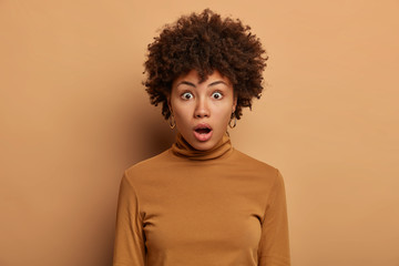 Fototapeta na wymiar Headshot of stunned woman with Afro hair, wonders something, gasps from excitement, keeps mouth opened, dressed in casual poloneck, isolated over beige background. No way. Unlikely to be true