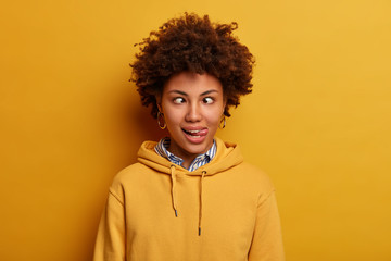 Photo of funny young woman has crazy face, crosses fingers and sticks out tongue, foolishes around,...