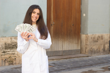 pretty caucasian young woman with dollar banknotes. Nurse, doctor, scientist, therapist, pharmacist, in the city, in white coat uniform, dark air. Place for your text in copy space.