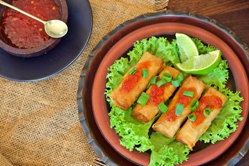 Spring rolls on a bed of green lettuce and two slices of lime next to a small bowl of sweet chilly sauce and a small spoon against a background of hessian