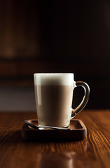 A clear glass mug filled with a creamy cappuccino or latte, topped with rich milk foam, placed on a wooden tray with a metal spoon beside it. 