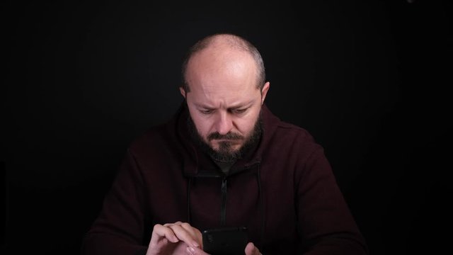 Adult caucasian man in hoodie sitting in front of black background, holding smart phone and typing