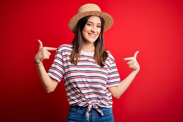 Young beautiful brunette woman wearing striped t-shirt and summer hat over red background looking confident with smile on face, pointing oneself with fingers proud and happy.