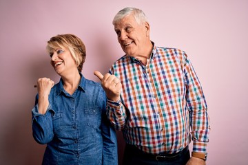 Senior beautiful couple standing together over isolated pink background smiling with happy face looking and pointing to the side with thumb up.