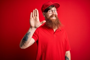 Naklejka premium Young handsome delivery man wearing glasses and red cap over isolated background Waiving saying hello happy and smiling, friendly welcome gesture