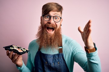 Redhead Irish cook man with beard holding maki sushi tray over isolated background very happy and excited, winner expression celebrating victory screaming with big smile and raised hands © Krakenimages.com