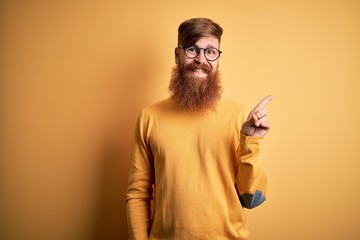 Handsome Irish redhead man with beard wearing glasses over yellow isolated background with a big smile on face, pointing with hand finger to the side looking at the camera.