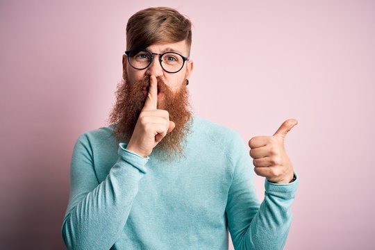 Handsome Irish redhead man with beard wearing glasses over pink isolated background asking to be quiet with finger on lips pointing with hand to the side. Silence and secret concept.