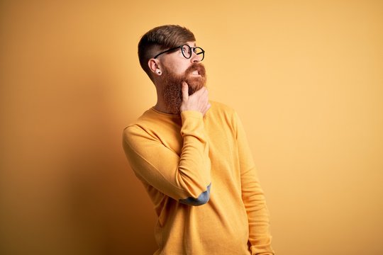 Handsome Irish redhead man with beard wearing glasses over yellow isolated background Thinking worried about a question, concerned and nervous with hand on chin