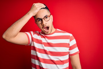 Young handsome man wearing casual striped t-shirt and glasses over isolated red background...