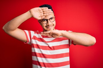 Young handsome man wearing casual striped t-shirt and glasses over isolated red background Smiling cheerful playing peek a boo with hands showing face. Surprised and exited