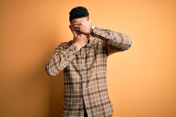 Young handsome man wearing casual shirt standing over isolated yellow background Covering eyes and mouth with hands, surprised and shocked. Hiding emotion
