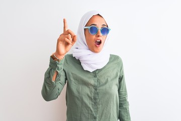 Young Arab woman wearing hijab and summer sunglasses over isolated background pointing finger up with successful idea. Exited and happy. Number one.