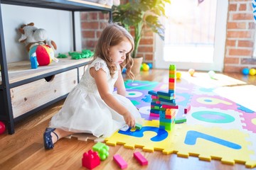 Adorable blonde toddler playing with building blocks toy around lots of toys at kindergarten