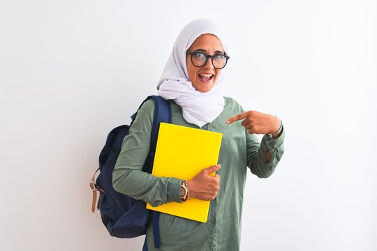 Young Arab student woman wearing hijab and backpack holding a book over isolated background very happy pointing with hand and finger