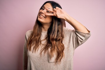 Naklejka premium Young beautiful brunette woman wearing casual sweater standing over pink background Doing peace symbol with fingers over face, smiling cheerful showing victory