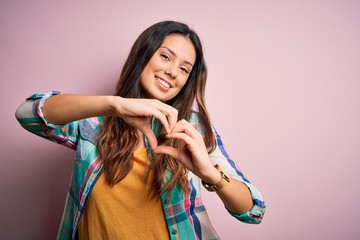 Young beautiful brunette woman wearing casual colorful shirt standing over pink background smiling in love doing heart symbol shape with hands. Romantic concept.