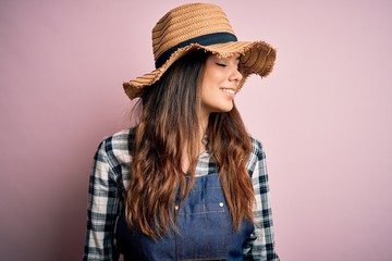 Young beautiful brunette farmer woman wearing apron and hat over pink background looking away to side with smile on face, natural expression. Laughing confident.