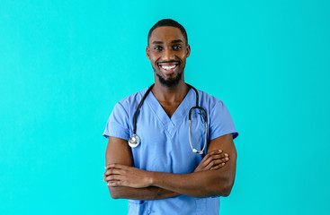 Portrait of a friendly male doctor or nurse wearing blue scrubs uniform and stethoscope, with arms crossed, isolated on blue studio background
