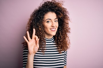 Young beautiful woman with curly hair and piercing wearing casual striped t-shirt showing and pointing up with fingers number three while smiling confident and happy.