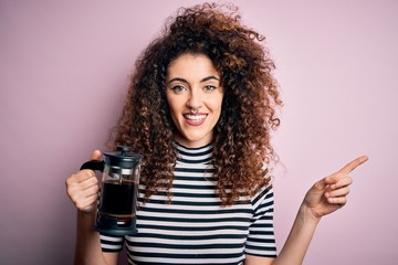 Young beautiful woman with curly hair and piercing doing coffee holding french coffeemaker very happy pointing with hand and finger to the side