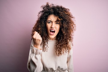 Beautiful woman with curly hair and piercing wearing casual sweater over pink background angry and mad raising fist frustrated and furious while shouting with anger. Rage and aggressive concept.