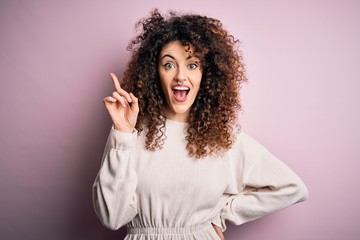 Beautiful woman with curly hair and piercing wearing casual sweater over pink background pointing finger up with successful idea. Exited and happy. Number one.