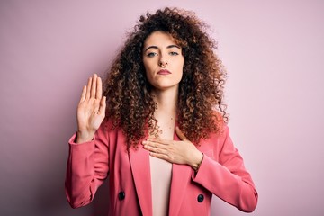 Young beautiful businesswoman with curly hair and piercing wearing elegant jacket Swearing with hand on chest and open palm, making a loyalty promise oath