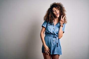 Young beautiful woman with curly hair and piercing wearing casual denim dress smiling with happy face winking at the camera doing victory sign. Number two.