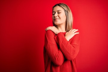 Young beautiful blonde woman wearing casual sweater over red isolated background Hugging oneself happy and positive, smiling confident. Self love and self care