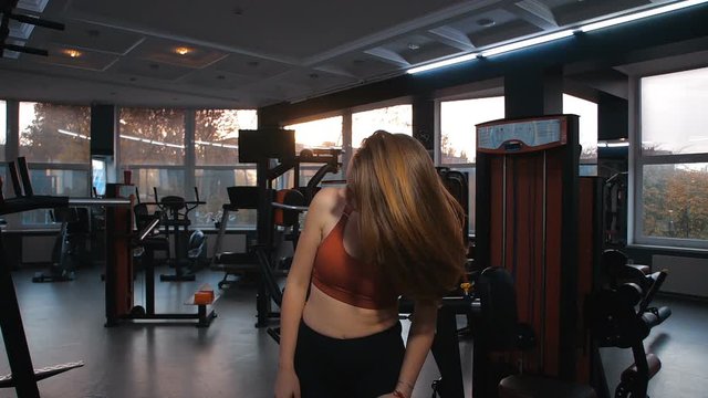 Female athlete in the gym. Portrait of attractive girl in black top and leggings standing and waving her  straight fair hair. Sunset in background.  200 FPS slow motion. Tracking shot, slide camera