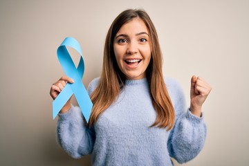Young blonde woman holding prostate cancer awareness campaing blue ribbon screaming proud and...