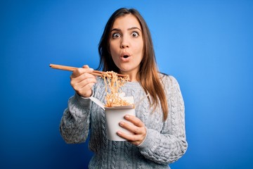 Young woman eating asian noodles from take away box using chopstick over blue background scared in shock with a surprise face, afraid and excited with fear expression