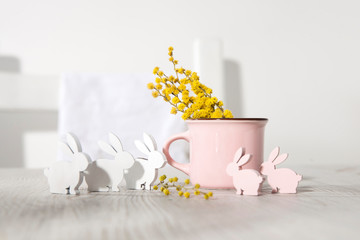 Pink ceramic milk mug with gypsophila and small plywood rabbits for decorating Easter table on a sunny morning