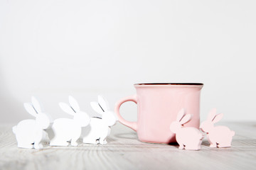 Pink ceramic milk mug with gypsophila and small plywood rabbits for decorating Easter table on a sunny morning