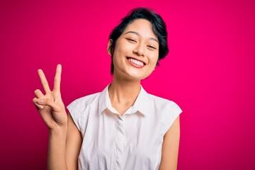 Young beautiful asian girl wearing casual summer shirt standing over isolated pink background smiling with happy face winking at the camera doing victory sign. Number two.
