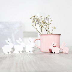 Pink ceramic milk mug with gypsophila and small plywood rabbits for decorating Easter table on a sunny morning