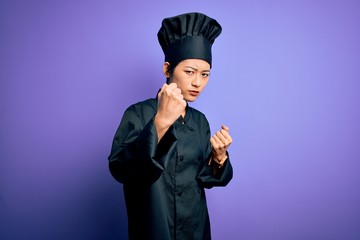 Young beautiful chinese chef woman wearing cooker uniform and hat over purple background Ready to fight with fist defense gesture, angry and upset face, afraid of problem