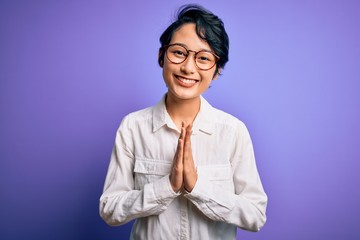 Young beautiful asian girl wearing casual shirt and glasses standing over purple background praying with hands together asking for forgiveness smiling confident.