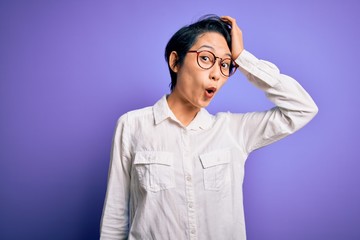 Young beautiful asian girl wearing casual shirt and glasses standing over purple background surprised with hand on head for mistake, remember error. Forgot, bad memory concept.