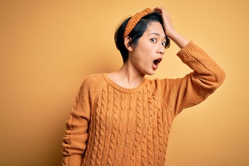 Young beautiful asian girl wearing casual sweater and diadem standing over yellow background surprised with hand on head for mistake, remember error. Forgot, bad memory concept.