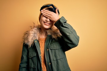 Beautiful asian skier girl wearing snow sportswear using ski goggles over yellow background smiling and laughing with hand on face covering eyes for surprise. Blind concept.