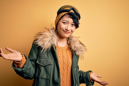 Beautiful asian skier girl wearing snow sportswear using ski goggles over yellow background clueless and confused expression with arms and hands raised. Doubt concept.