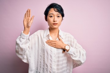 Young beautiful asian girl wearing casual shirt standing over isolated pink background Swearing with hand on chest and open palm, making a loyalty promise oath